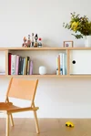 A photo of a white Nest WiFi router on a shelf next to books. It blends in with the decor easily. There is a wooden chair sitting in the room in front of the shelf. There is also a vase with flowers a photograph on the shelf.