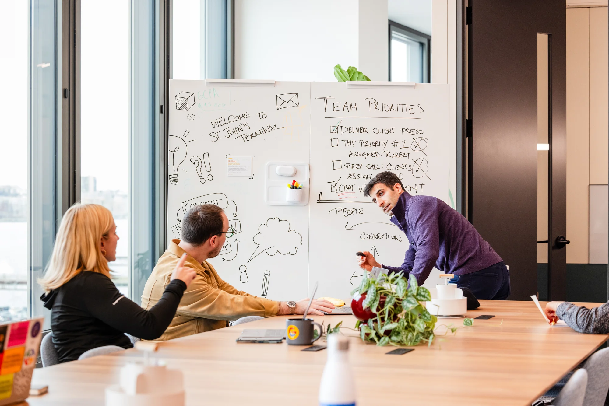 A Googler in a purple sweater stands at a whiteboard, collaborating with two of his colleagues seated at a communal table.