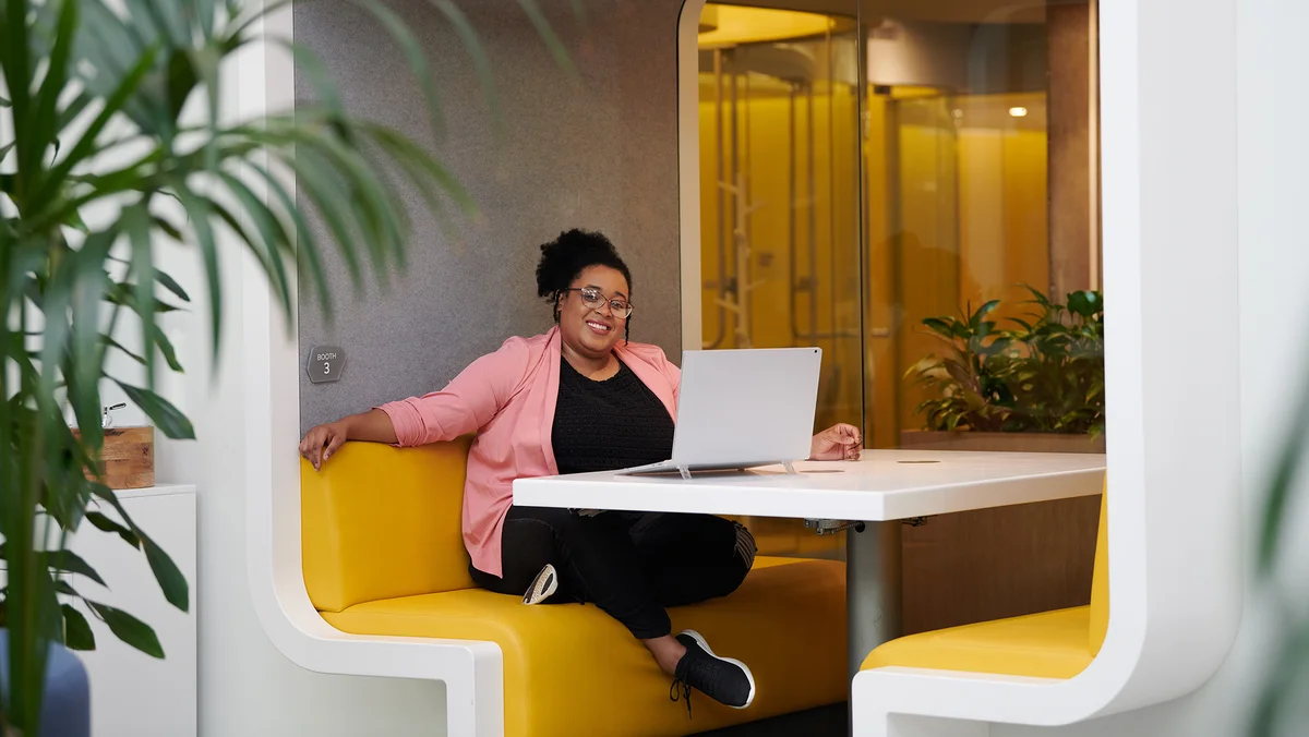 A woman sits in a booth with a laptop, turning to the camera and smiling.