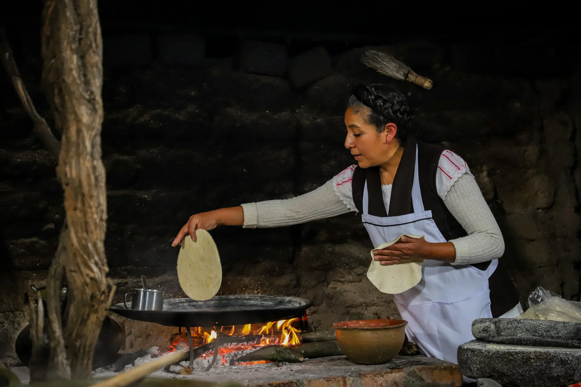 A woman wearing an apron putting a tortilla on a pan over an open flame