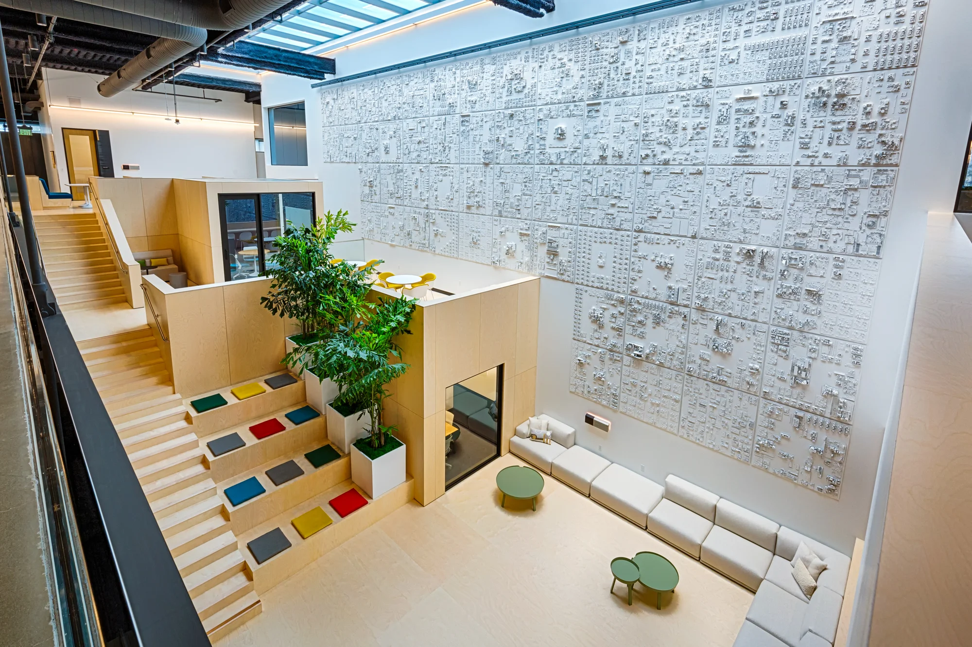 Inside the building there are multiple levels of seating, including amphitheater seating, a large sectional couch and small tables with chairs. The space is open and airy and punctuated with colored cushions and large potted plants. One of the walls is covered in white paneling made out of circuit boards.