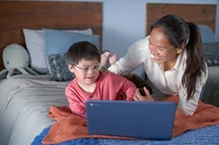 Image of woman and her young son who wears glasses lay on his bed using a Chromebook for schoolwork.