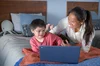 A young boy wearing glasses is lying on a bed looking at a Chromebook, with his mother next to him.