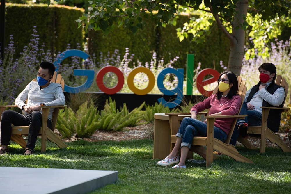 Three masked people sit near a “Google” sign in adirondack chairs on a lawn.