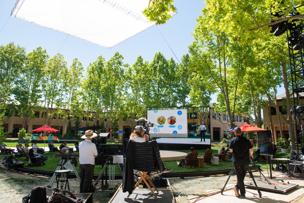 A camera crew of several people are in the foreground, filming a stage surrounded by trees.