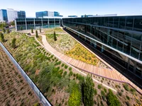 An aerial shot of a building shows an L-shaped office building with large glass windows. Outside the landscaping is made up of green shrubbery and bushes with yellow flowers spotted with coastal live oaks and divided by a paved walking path.