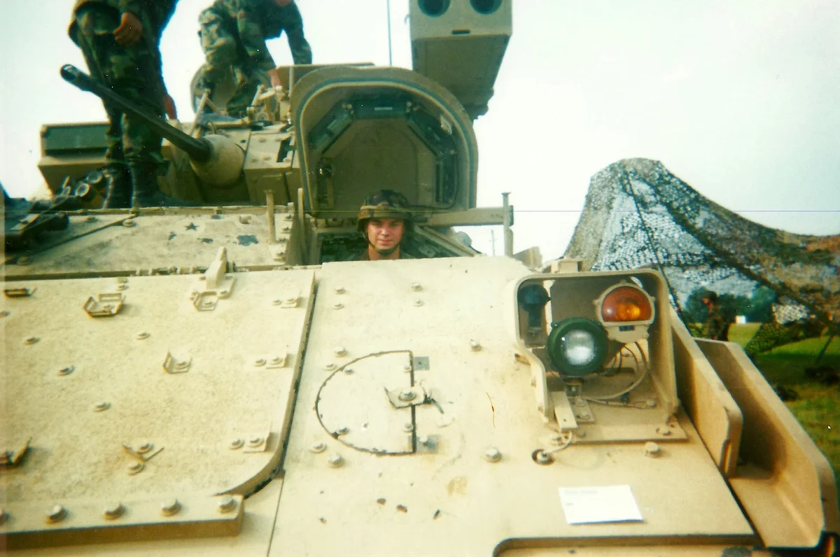 Dennis James smiling and sitting in a tan military vehicle called a Bradley Fighting Vehicle. Two other soldiers are pictured in the top-left corner standing on top of the vehicle. le's hero media