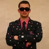 Shea standing indoors wearing sunglasses and a suit decorated with pac man figurines.