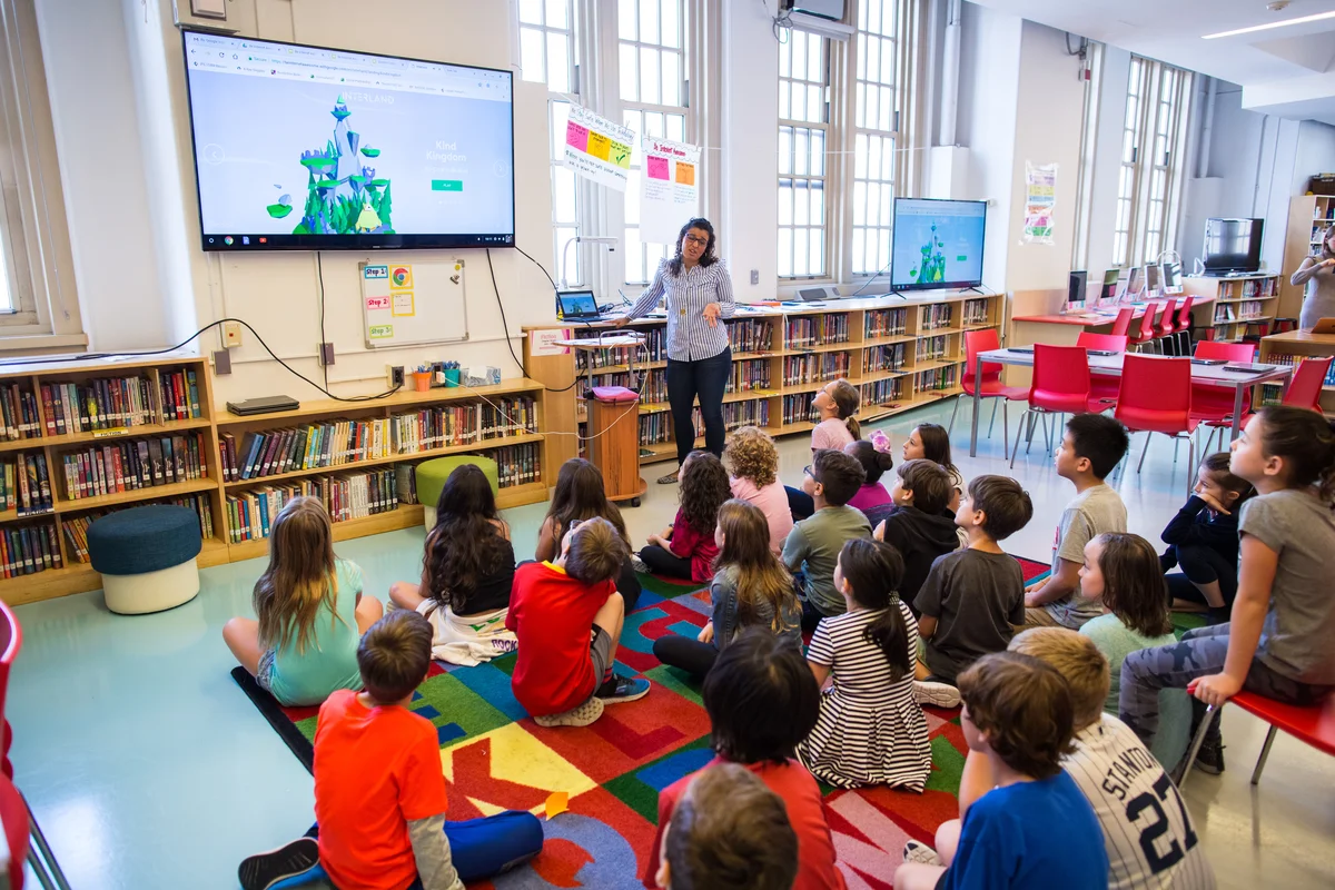 Nauczyciel w okularach i koszuli w niebieskie paski prowadzący prezentację wideo dla grupy uczniów szkół podstawowych. Dzieci siedzą na kolorowym dywanie w szkolnej bibliotece.