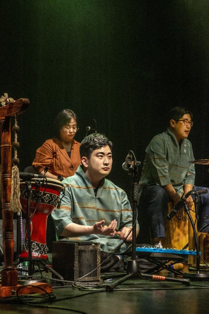 Seokhyun Lee wearing a traditional Korean outfit (in green), sitting on a stage surrounded by musical instruments and microphones. Behind him are two other members of the ensemble, one a woman over his right shoulder (wearing yellow), one behind him and to his left (wearing the same green outfit as Seokhyun).