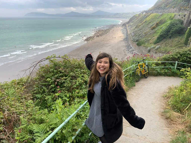 Desi steht auf einem Weg, lächelt in die Kamera und zeigt mit ihrer rechten Hand auf den Strand von Bray in Irland hinter sich.