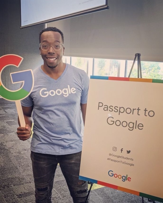 Brandon, wearing glasses and a light blue Google t-shirt, smiles and holds up a cutout of the Google logo while standing next to a sign that says “Passport to Google.”