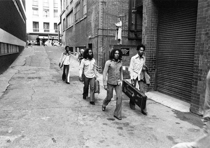 Bob Marley and three other men holding instrument cases walking down a street