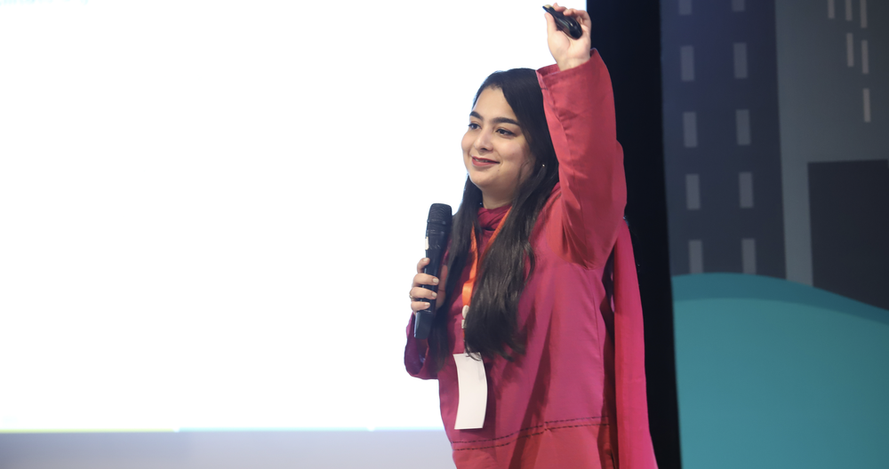 Photograph of a woman looking out in to a crowd smiling with her arm raised in the air. She is wearing a purple tunic and holding a microphone. Her name tag reads "Hufsa."