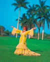 Singer Celia Cruz standing on grass wearing a yellow gown, arms outstretched and singing.