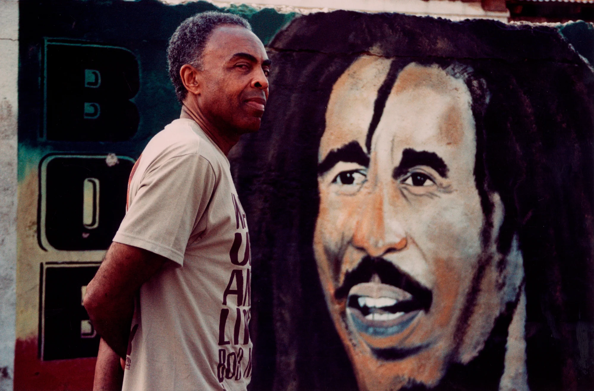 Gilberto Gil posing in front of a mural with the image of Jamaican singer Bob Marley