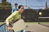 Kobieta na wózku inwalidzkim gra w pickleball na zewnętrznym betonowym boisku. Jej partnerka do gry podwójnej w niebieskiej koszulce stoi przy siatce i obserwuje, jak przygotowuje się do uderzenia.