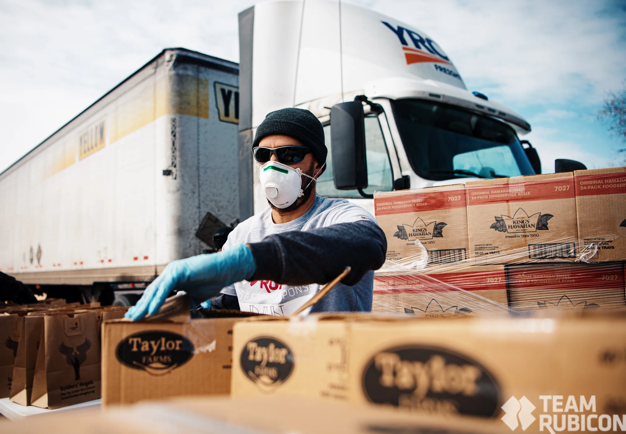 A volunteer wearing mask organizes a pile of boxes in front of a semi truck.
