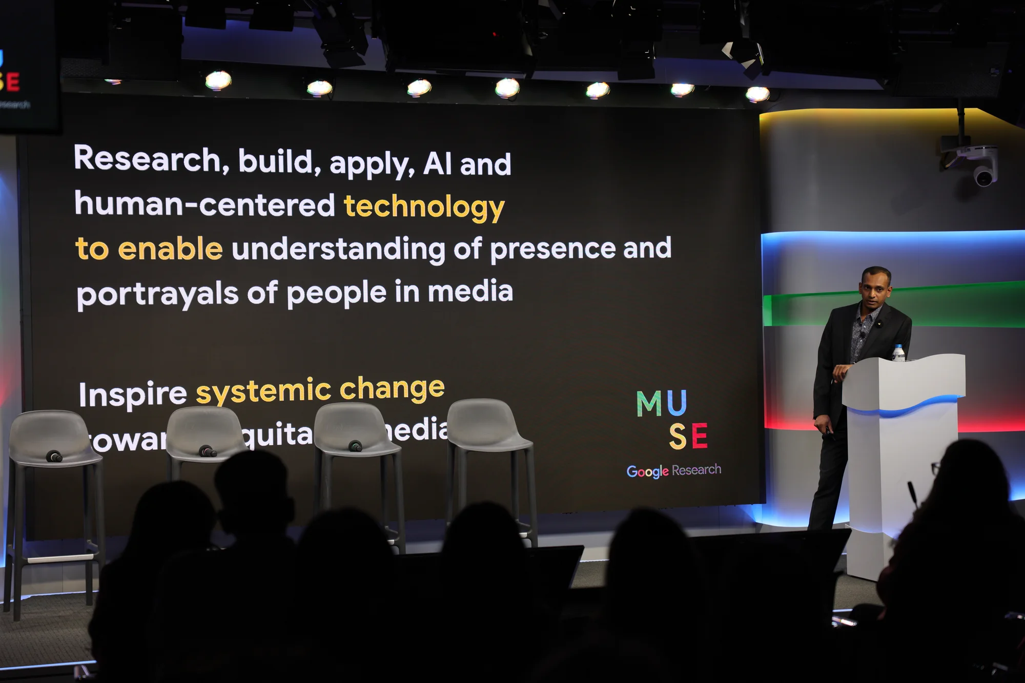 Person standing in front of a wall-sized presentation screen. Presentation showing text: “Research, build, apply, AI and human-centered technology to enable understanding of presence and portrayal of people in media. Inspire systemic change towards equitable media.”