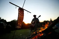A man playing a guitar sits next to a rack of beef roasting over an open fire.