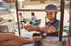 Ciemnoskóra osoba ubrana w kask rowerowy i smartwatch uśmiecha się, z ręką wyciągniętą nad terminalem POS przy oknie food trucka. Po lewej stronie napis: „G Pay”. Za nimi ludzie siedzą przy stoliku z kawą i rozmawiają.