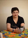 South Asian woman wearing a black shirt, white smartwatch on her right hand, and grey prosthetic as her left forearm. She smiles at the camera ​​and rests her hands on a colorful table.