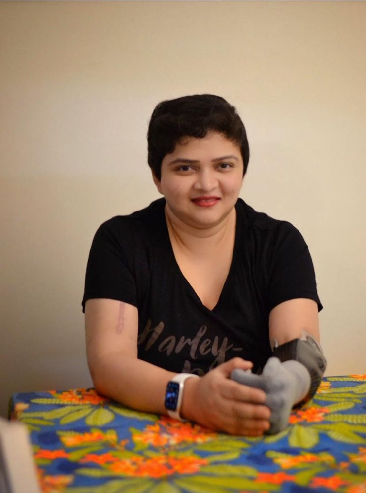 South Asian woman wearing a black shirt, white smartwatch on her right hand, and grey prosthetic as her left forearm. She smiles at the camera ​​and rests her hands on a colorful table.