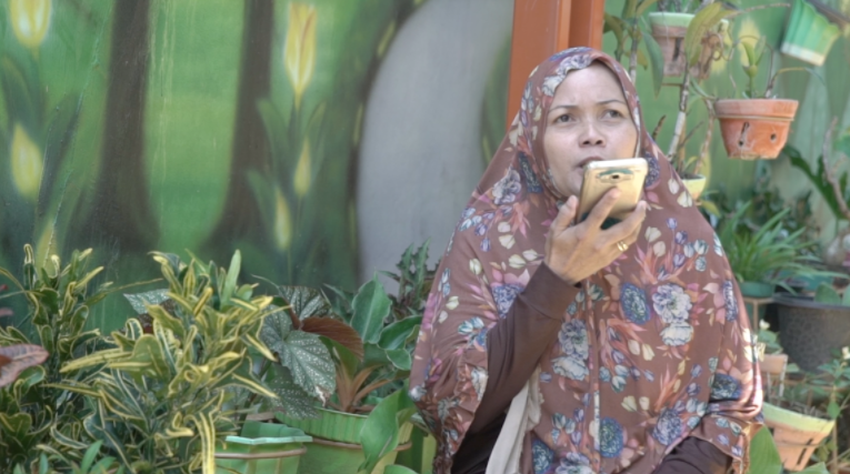 A woman in traditional dress in Indonesia sits facing the camera and talking to her phone. She’s surrounded by potted plants.