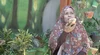 A woman in traditional dress in Indonesia sits facing the camera and talking to her phone. She’s surrounded by potted plants.