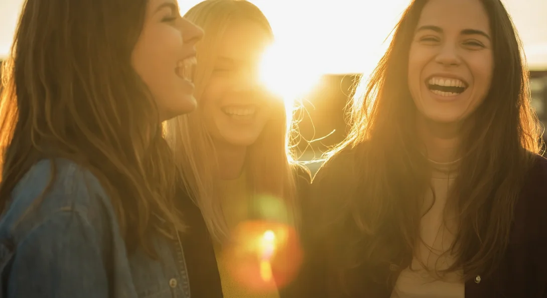 Prompt: Three women stand together laughing, with one woman slightly out of focus in the foreground. The sun is setting behind the women, creating a lens flare and a warm glow that highlights their hair and creates a bokeh effect in the background. The photography style is candid and captures a genuine moment of connection and happiness between friends. The warm light of golden hour lends a nostalgic and intimate feel to the image.