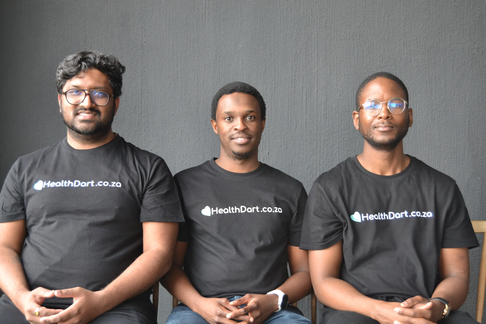 Three men wearing black t-shirts with "Healthdart" written in white letters all face the camera, sitting with their hands folded.