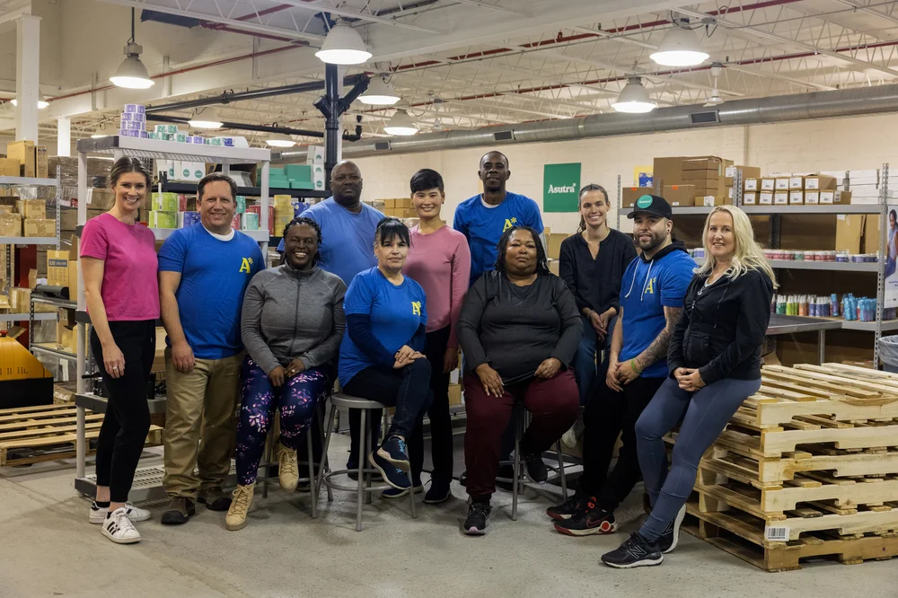 A part of the team at Austra, sitting and standing in a warehouse-like room with shelves stocked with boxes and wood palettes scattered around. They are in a tight group smiling at the camera.
