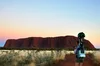 A woman with a camera attached to her backpack looks towards Uluru in the distance, as she films footage for Street View. The sun is setting behind Uluru.