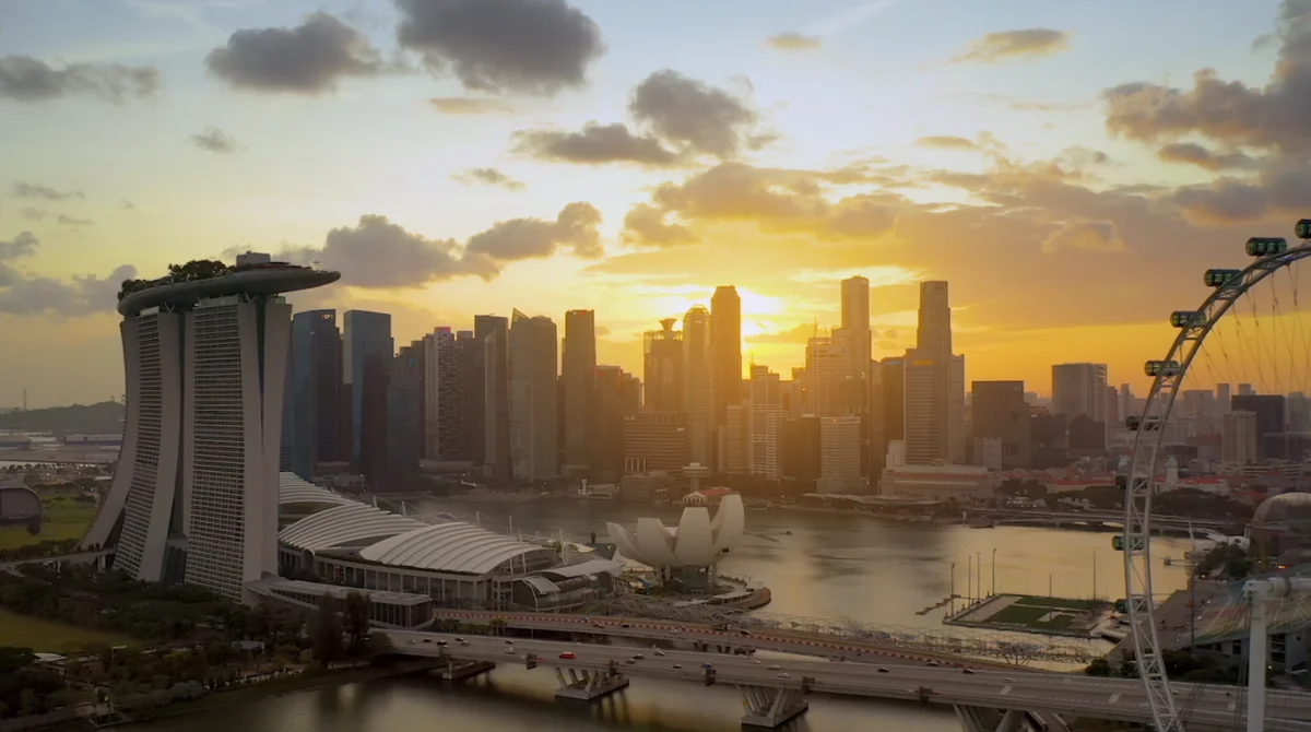An image of the Singapore skyline with the sun setting behind scattered clouds in the background. There are skyscrapers in the centre of the photo, the glassy water of Marina Bay in the center, and an obvervation wheel to the right.