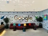 Ein Empfangsbereich im Büro von Google Dubai mit einer roten Couch, drei schwarzen Beistelltischen und dem in Kacheln geschriebenen Wort „Google“ an einer wellenförmig aussehenden Wand.