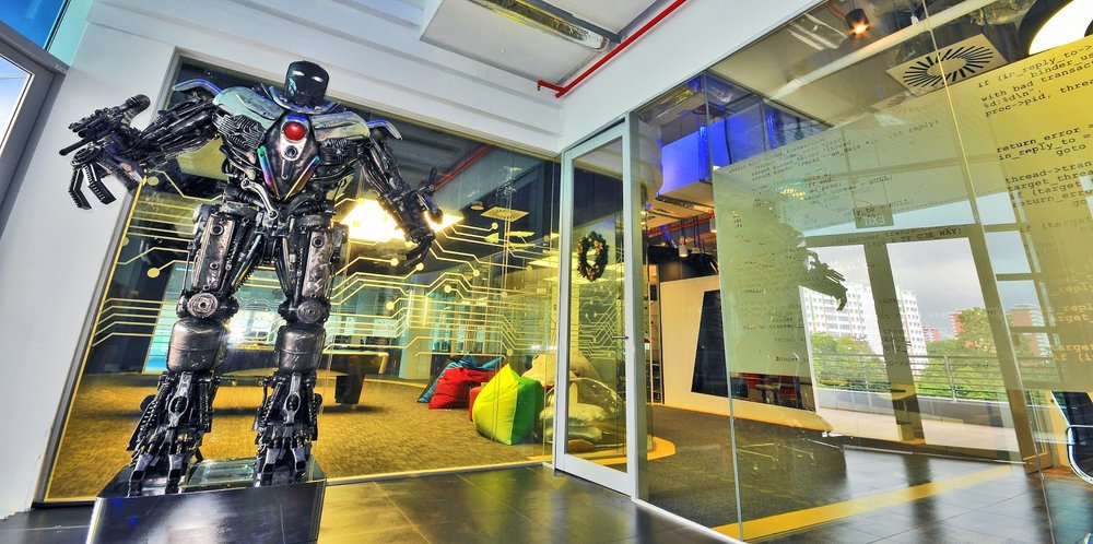 A tall robot-like figurine standing on a plinth in front of the entrance to one of Google’s data centers in Singapore. Beanbags and a pool table are visible through the glass office walls.