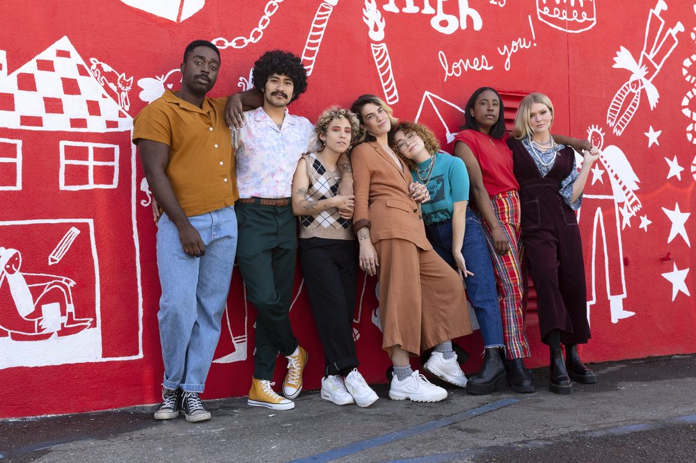 A group of seven racially diverse and gender expansive people stand together lovingly in front of a red and white mural at Junior High.