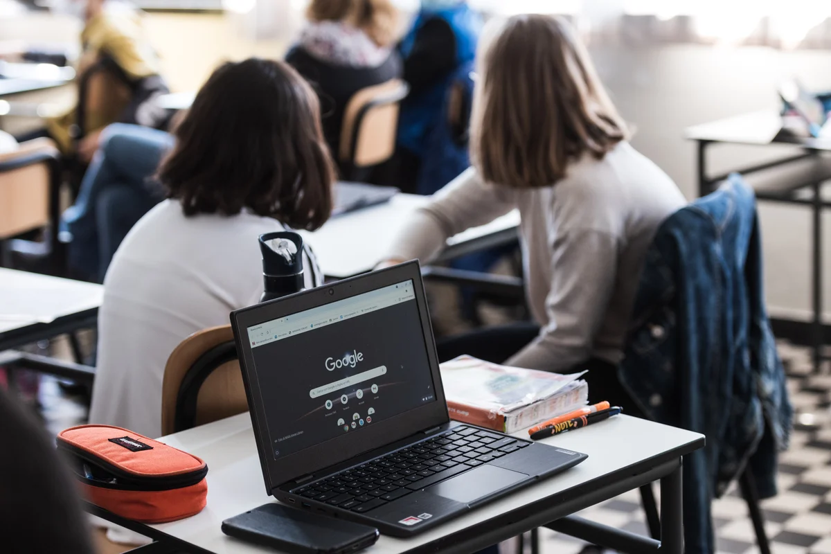 An image of a laptop on a classroom table with Google search on the screen. Two school girls sit in the background with their backs to us.