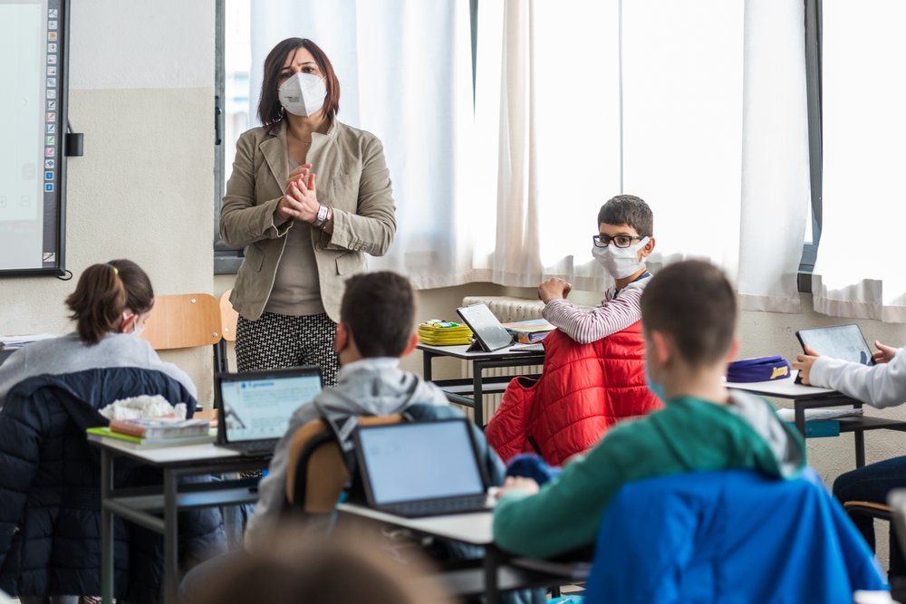 A picture of a teacher wearing a mask teaching children in a classroom who all have their laptops open