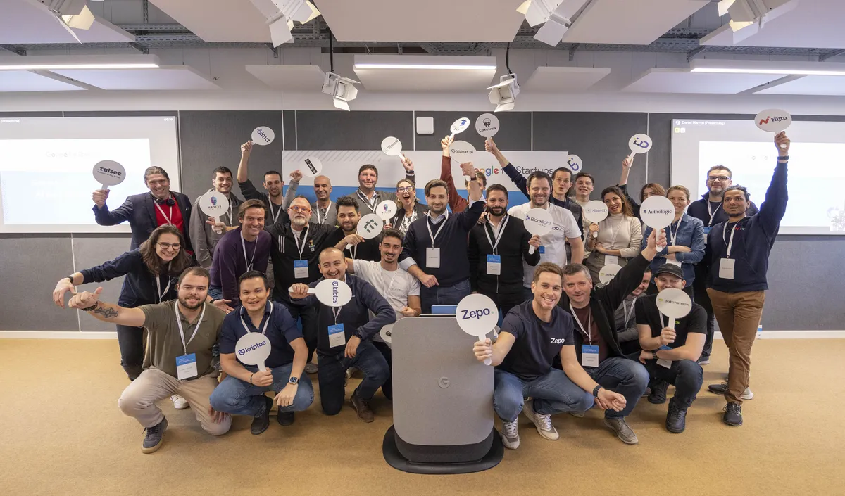 A group of people stand together in an office, some crouching, holding white round signs with their startup logos.