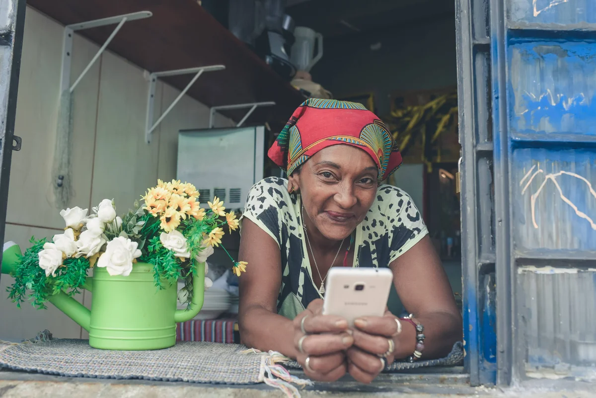 A woman with her hair wrapped up in a red scarf is smiling as she holds a smartphone in both hands and perches on a window sill.