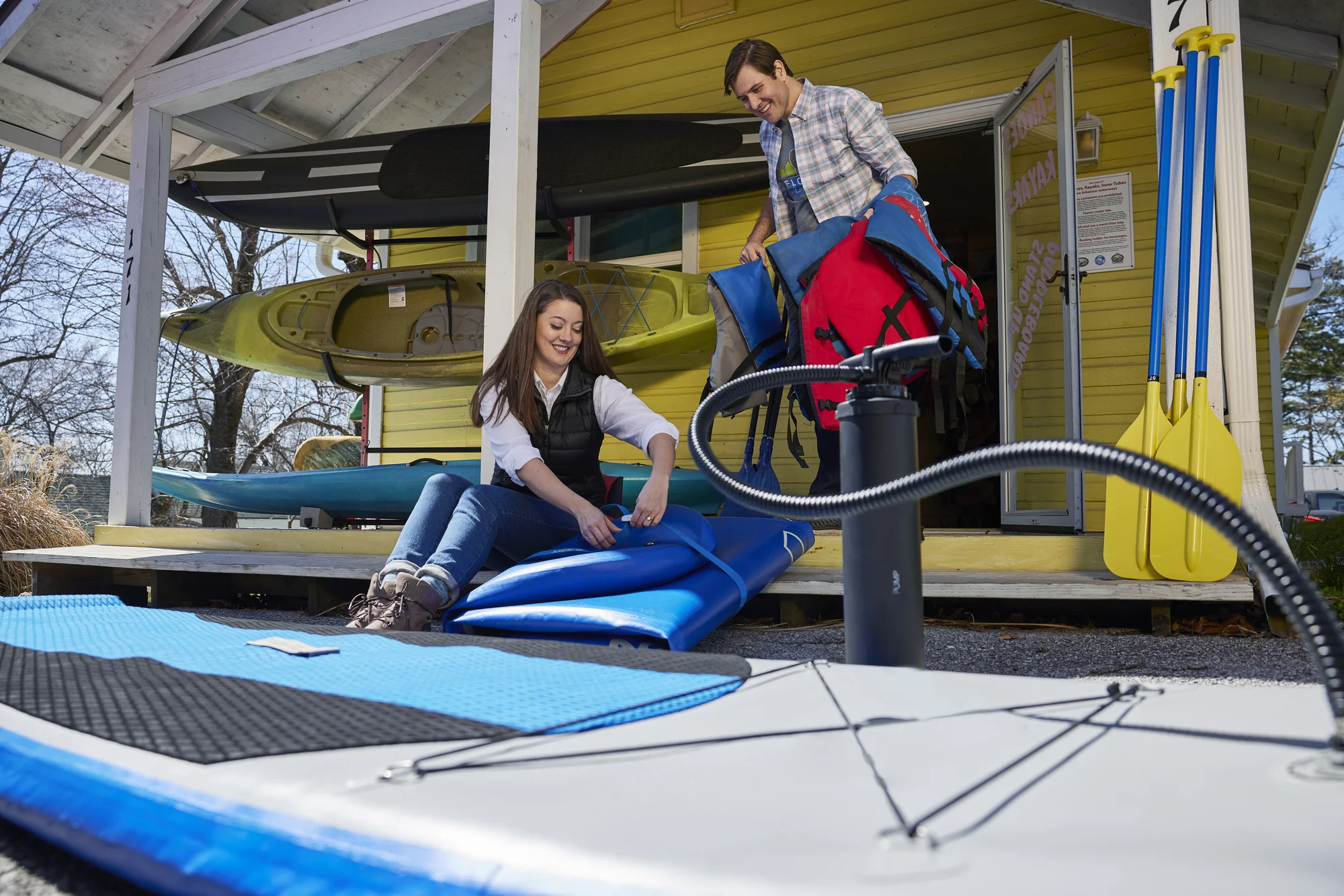 Owners Max (standing) and Rachel (sitting) outside a bright yellow boatshed with kayaks attached to the front. Both are holding safety equipment.