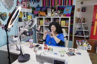 Owner Kathy at her workshop desk surrounded by colorful craft materials. She’s smiling and holding up one of her creations for a smartphone set up to record.