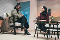 Zwei Frauen sitzen gegenüber in gepolsterten Stühlen, Adanna lacht, beiges Jacket und dunkle Hose, Alaa in weinroter Jacke und dunkler Hose sowie Kopftuch