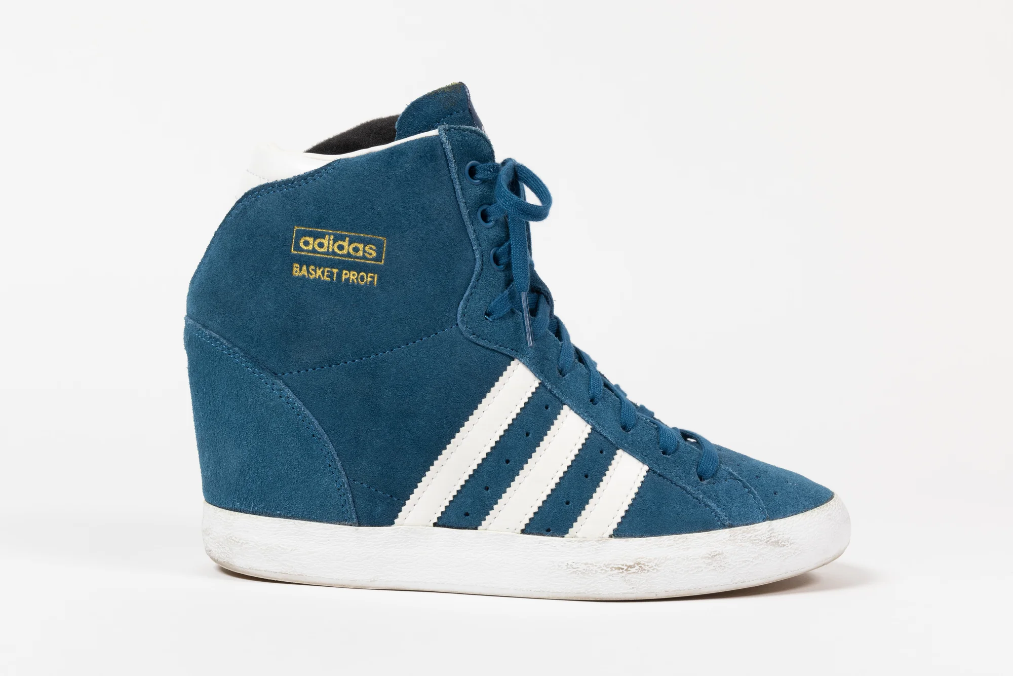 A color photograph of a turquoise Adidas wedge sneaker. The upper is constructed with a combination of suede and synthetic materials in turquoise, featuring the iconic Adidas three stripes on the sides in a contrasting white color. The sneaker sports a wedge and the Adidas logo on the side in gold.