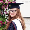 Image shows a young woman with long red hair and glasses looking over her shoulder and smiling at the camera. She's wearing a graduation cap and gown and standing in front of a wall of flowers.