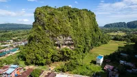 an aerial view of a tall rock that is covered in trees and green foliage, with an opening in the center to the cave site. There are a few buildings surrounding it.
