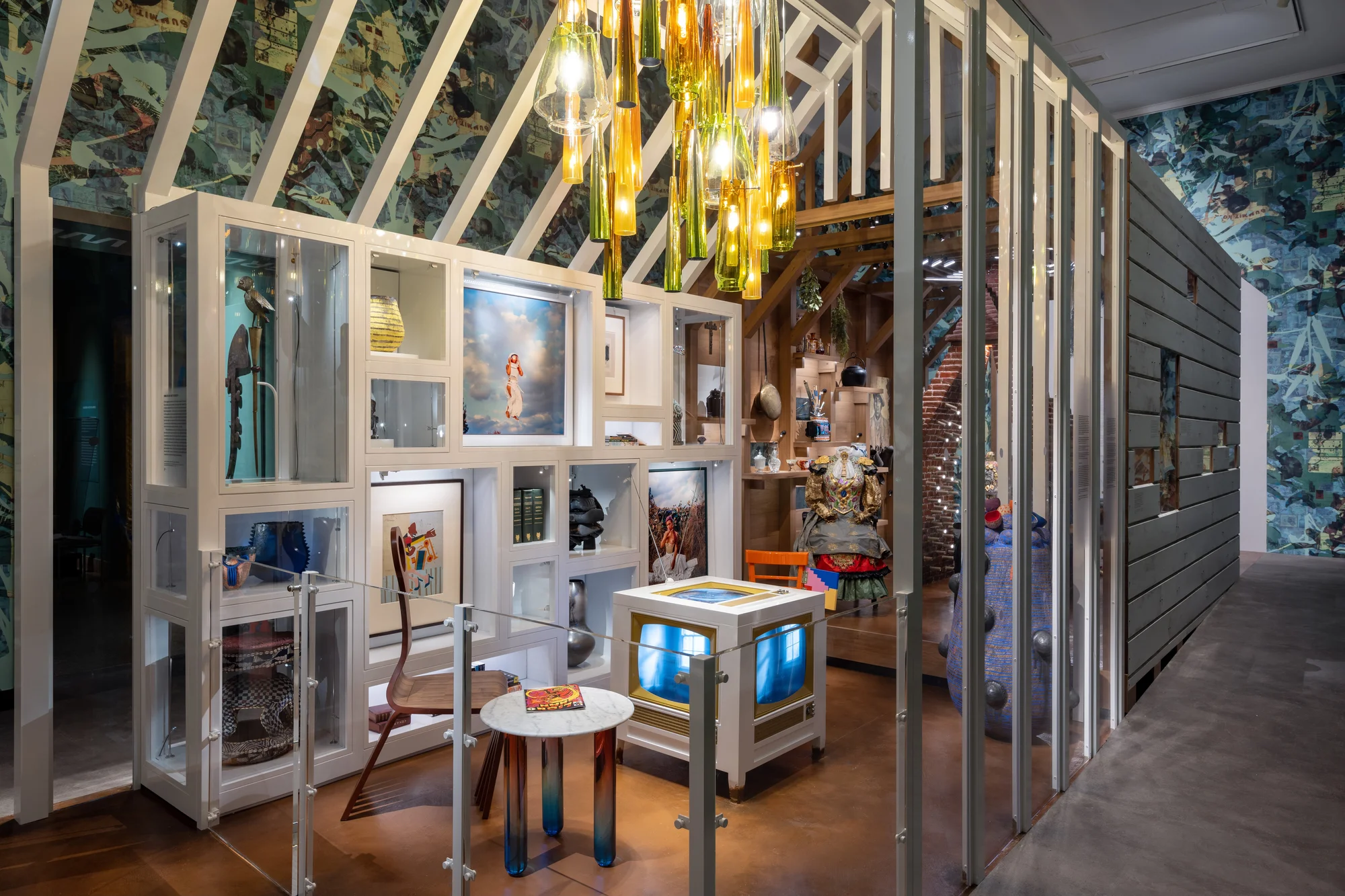 An image of an exhibition installation of an home’s interior; a green and yellow glass chandelier, a bookcase with each shelf filled with a different artwork or artifact, and living room with a chair with legs like a comb and a table and television at the center of the room.