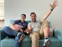 Two people sit together on a blue sofa with pink pillows. They are smiling and waving their hands in the air.