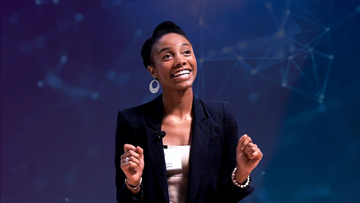 Sarah Murphy Gray is wearing a black blazer and white blouse, smiling broadly and looking out into the audience as she gestures with her hands during a talk at an AI conference.